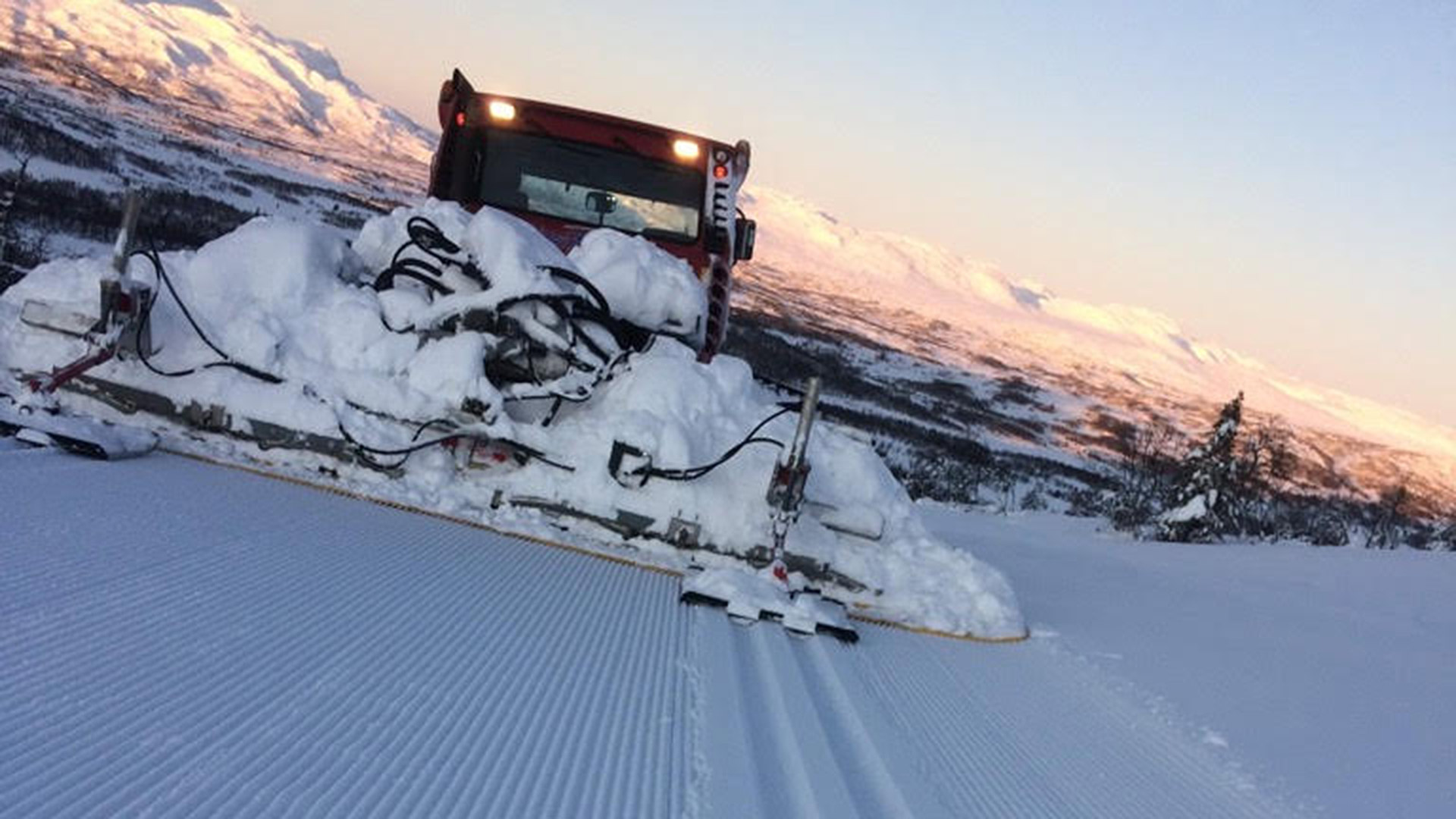 Snøplog som preparerer løyper i vinterlandskap ved solnedgang i fjellene.