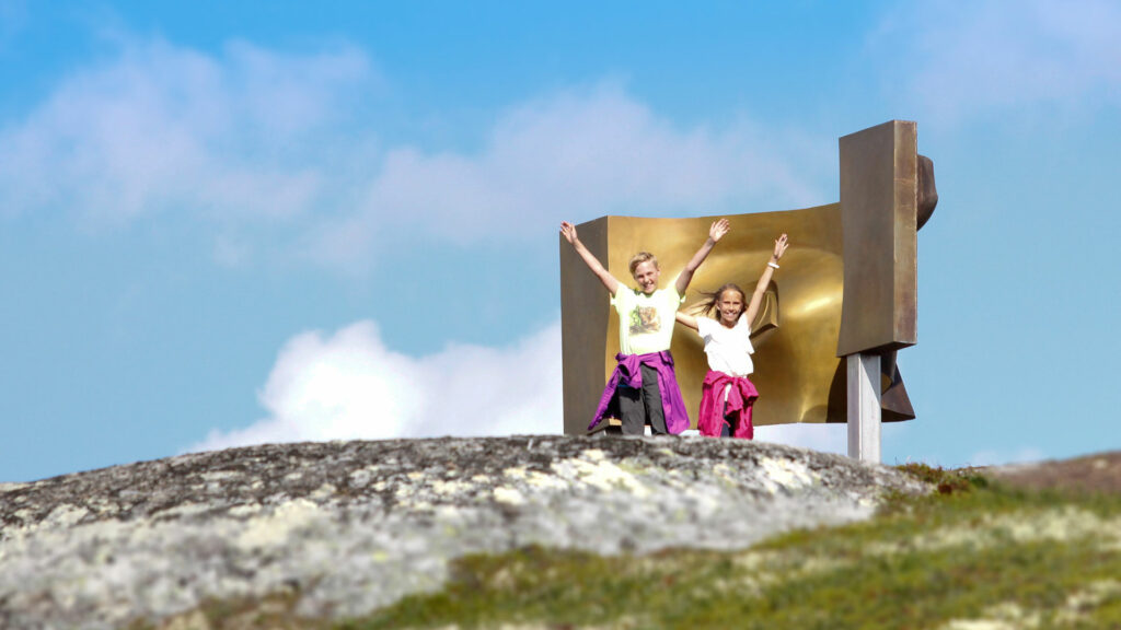 To barn jublende på fjelltopp med kunstinstallasjon i bakgrunnen, blå himmel og skyer, sommerstemning.