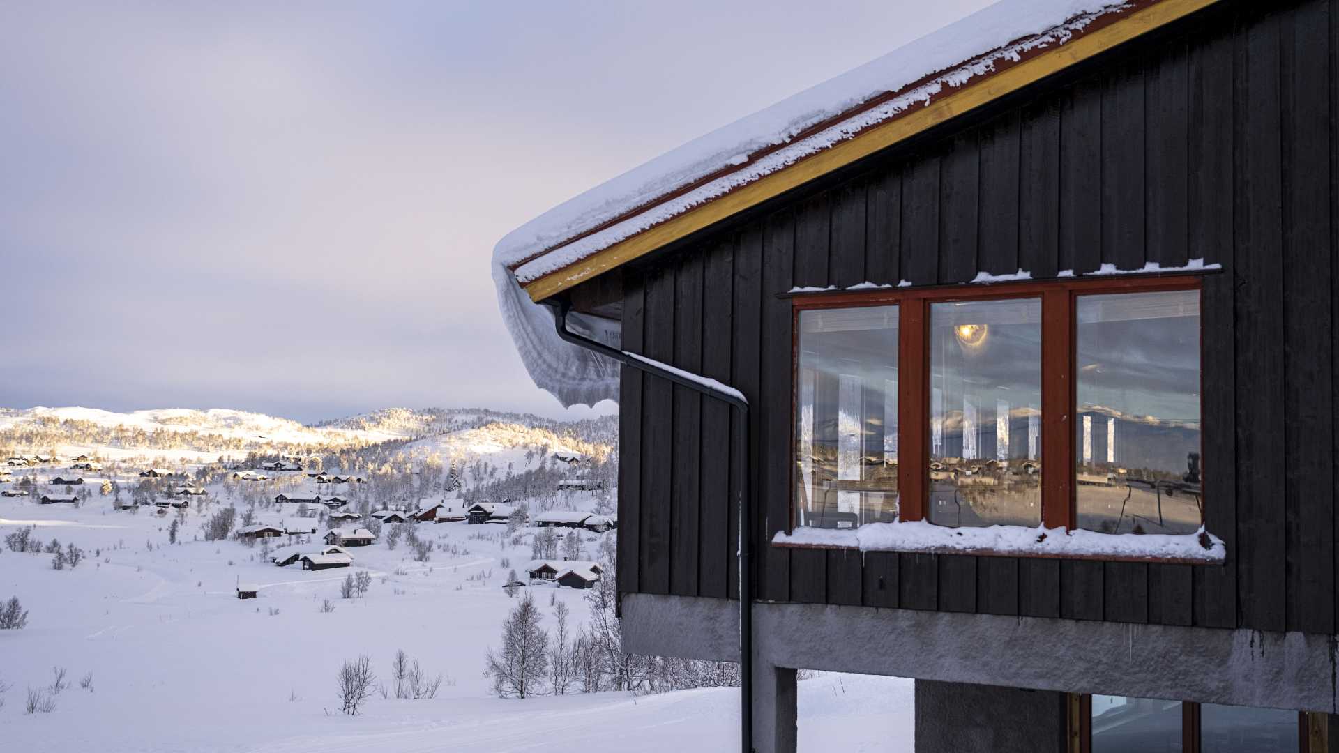 Schwarzes Holzhaus in einem schneebedeckten norwegischen Dorf, mit Blick auf sonnenbeschienene Berge am Horizont.