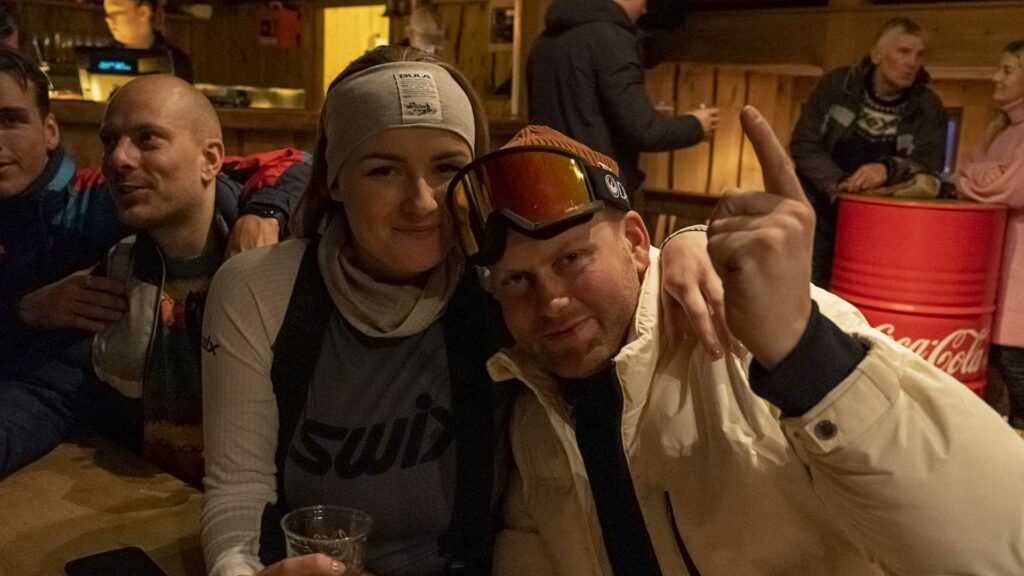 Personer på après-ski-bar, smiler og koser seg med drikke, vinterstemning innendørs.