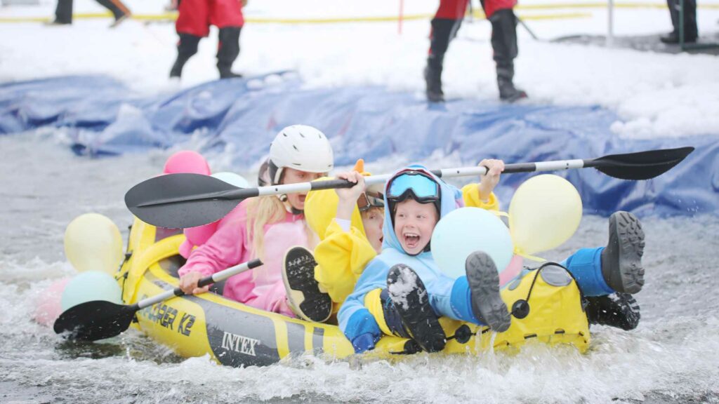 Barn i fargerike kjeledresser har det moro i gummibåt på vannet om vinteren. Ballonger festet.
