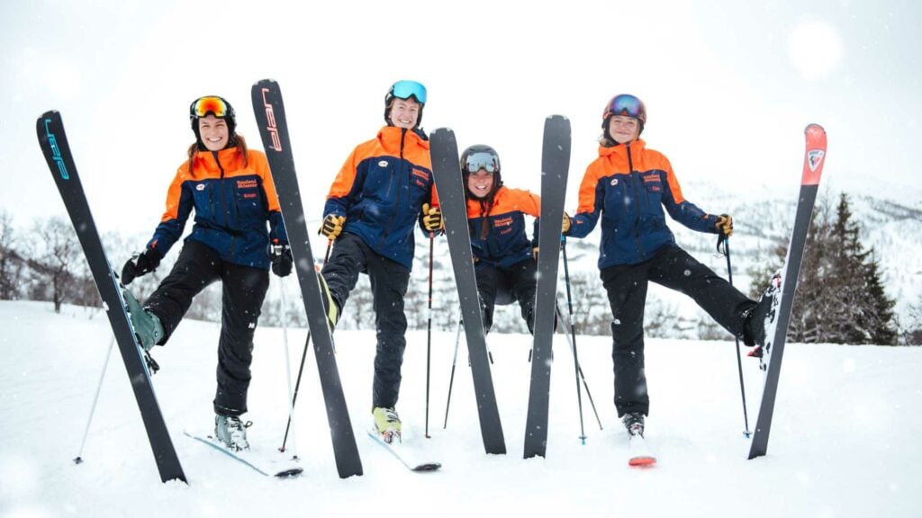 Fire glade skilærere poserer på ski i snødekte fjell med blå og oransje jakker.