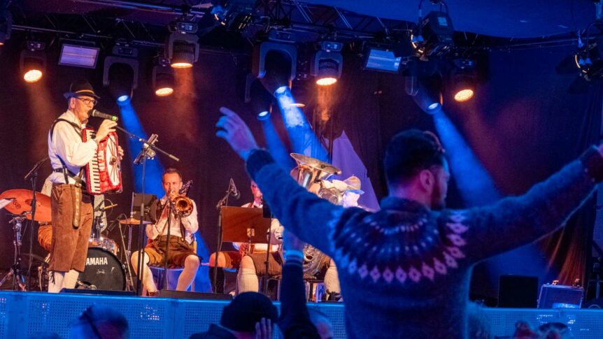 Musikere i tradisjonelle klær spiller på en scene, sett fra bak en entusiastisk publikummer.