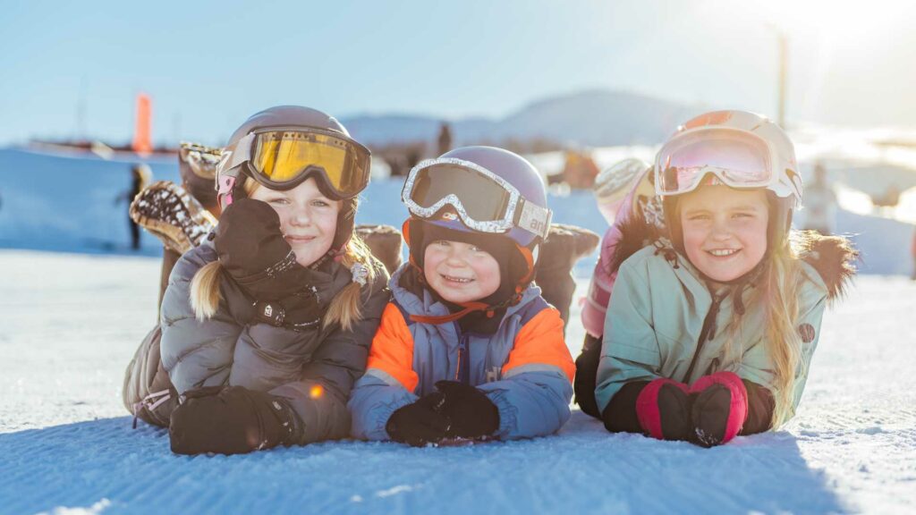Tre barn med skiutstyr smiler på snødekt bakke i solskinn. Vintermoro og utendørsaktivitet for barn.