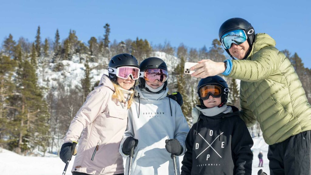 Familie på ski tar selfie i solfylt vinterlandskap med trær i bakgrunnen.