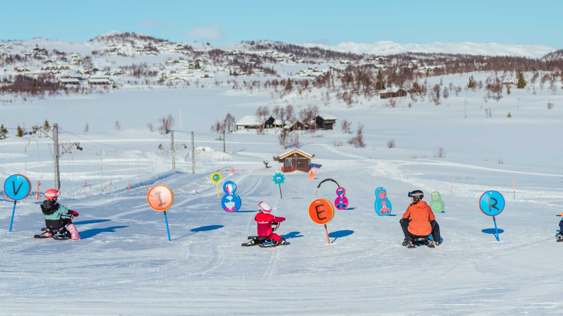 Barn som aker på en snødekt bakke med fargerike skilt og figurer i vinterlandskap.