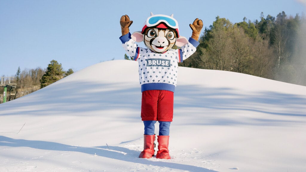Maskot i vinterklær jubler på snødekt område med skog i bakgrunnen.