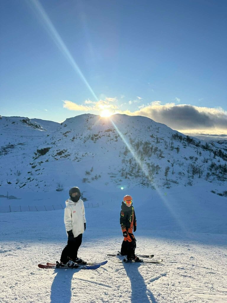 To personer står på ski i solfylt fjellandskap. Klar himmel og snødekte topper i bakgrunnen.