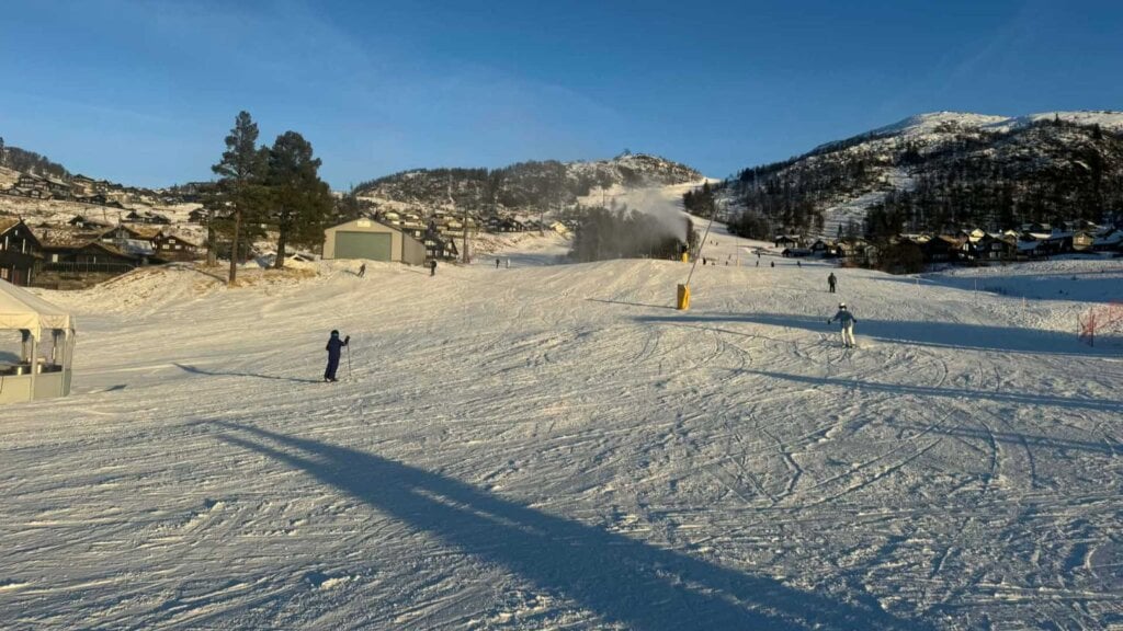 Skianlegg med mennesker, hytter og snøkanon i fjellene under blå himmel i Norge.