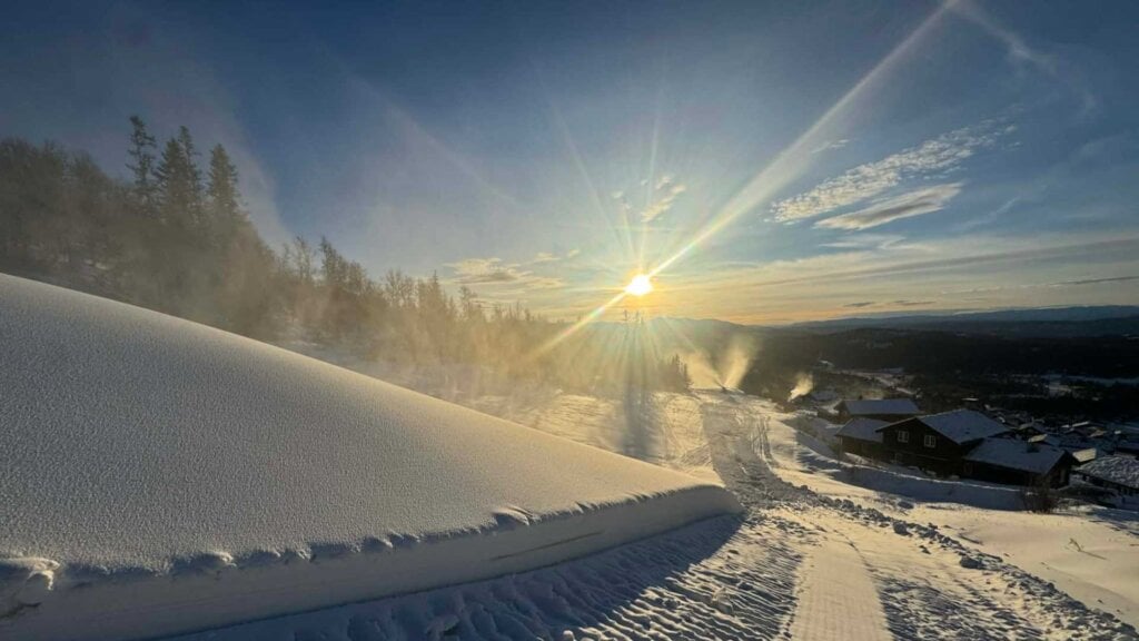 Solnedgang over snødekt landskap med hus, trær og strålende sollys i det fjerne.
