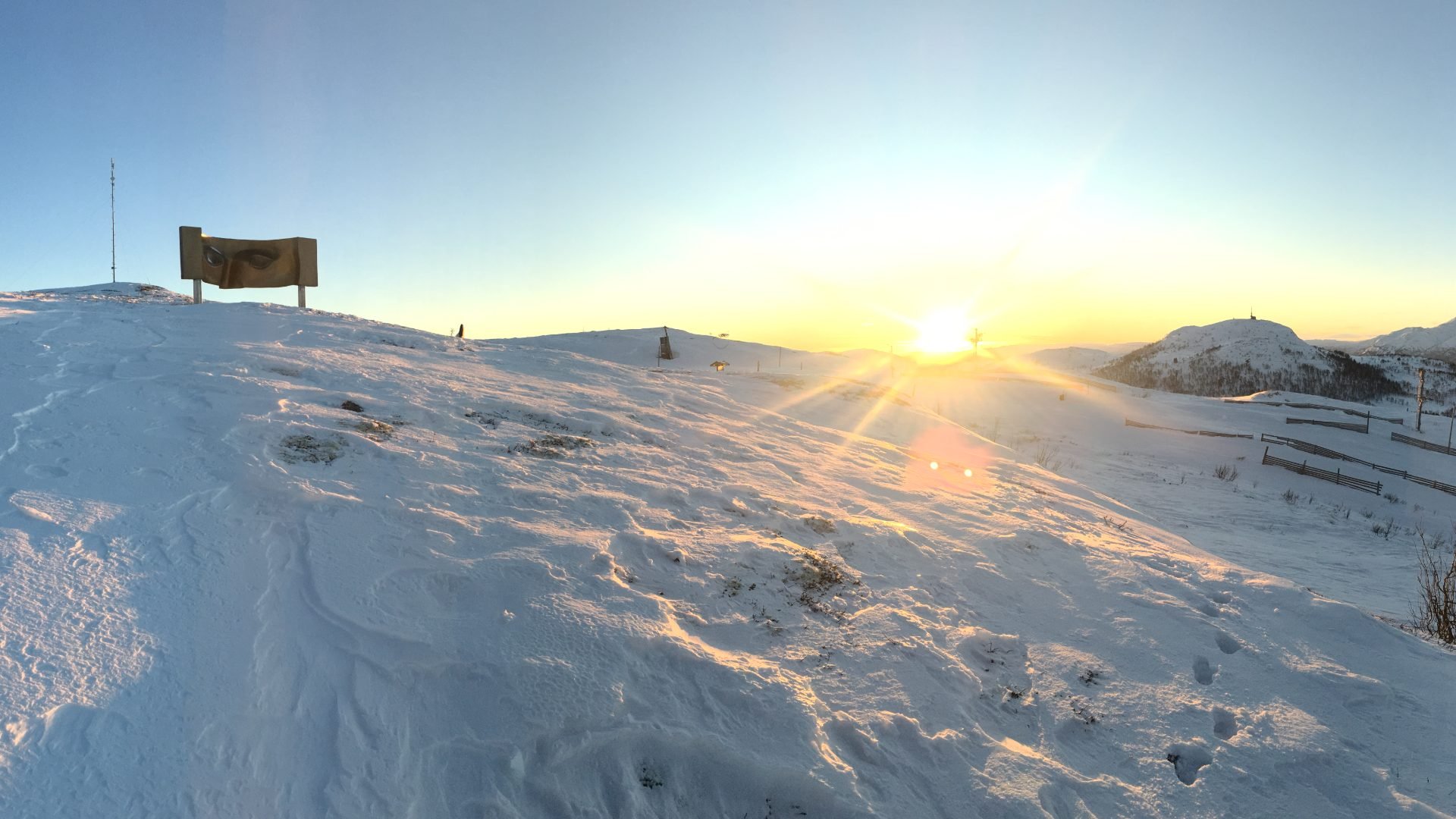 Vinterlandskap med snøkledde fjell i solnedgang, en stor skulptur og klar blå himmel.