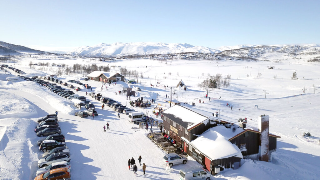 Vinterlandskap med skiløpere, parkerte biler og hytter i solfylt fjellområde.