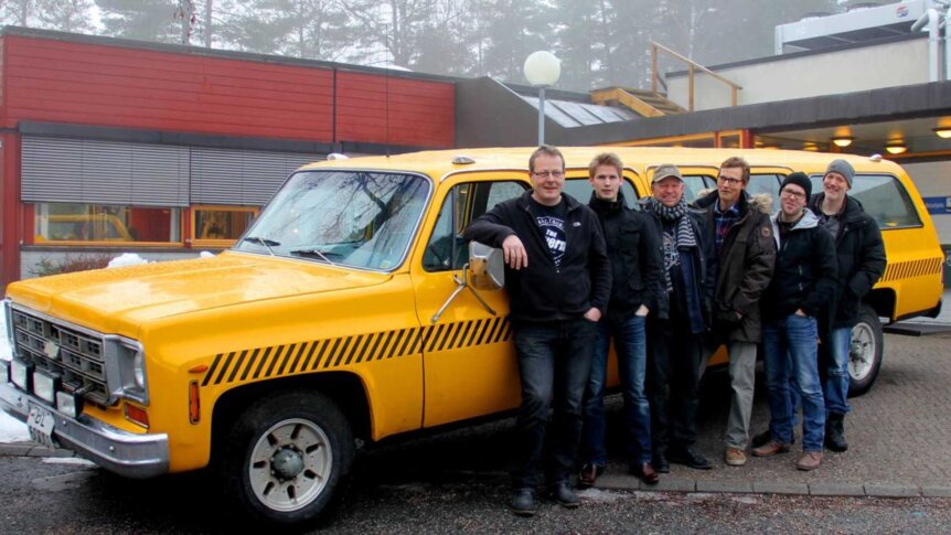 En gruppe menn poserer foran en gul vintage bil utenfor en bygning i vintermiljø.