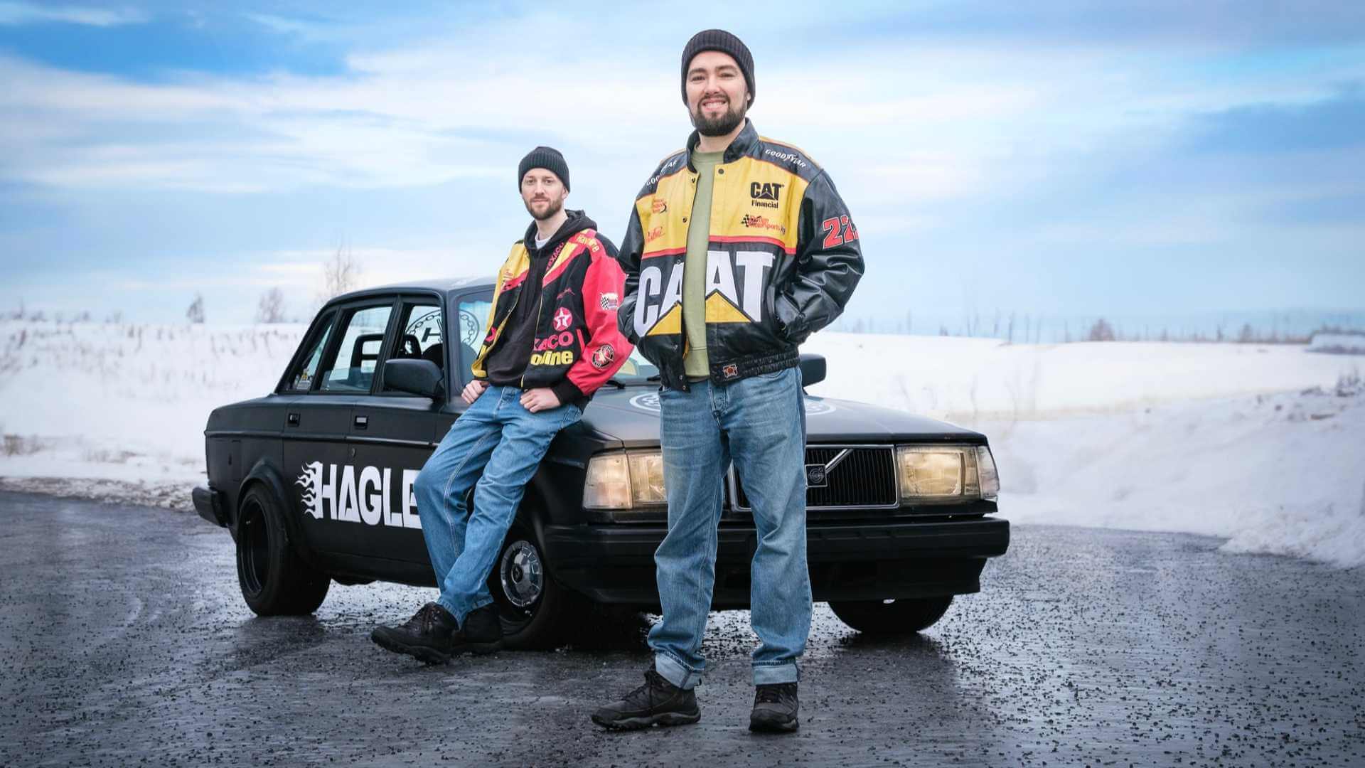 To personer i fargerike jakker poserer ved en svart bil om vinteren.