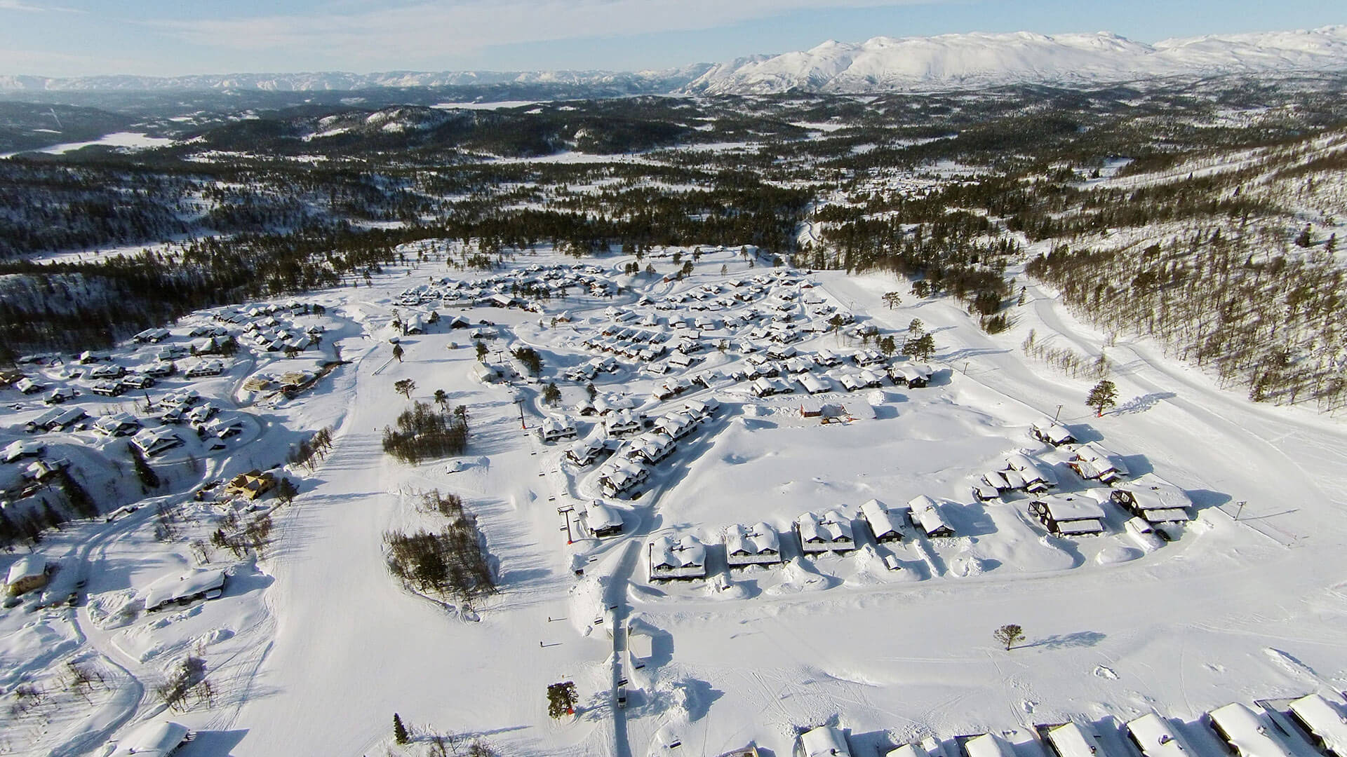 Luftfoto av snødekt norsk fjellandsby med fjell i bakgrunnen, klare vinterforhold.
