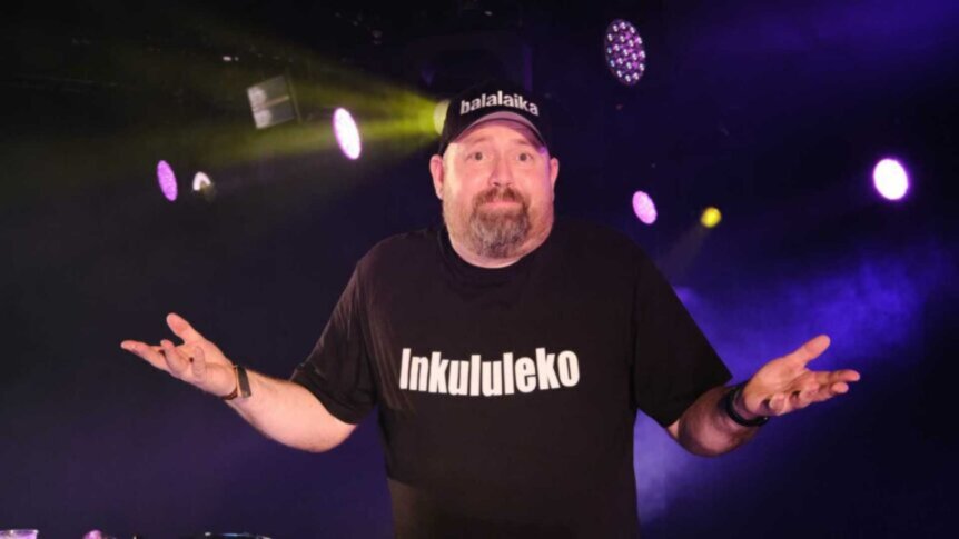 Mann på scenen med t-skjorte som sier Inkululeko og caps, i fargerikt opplyst konsertmiljø.