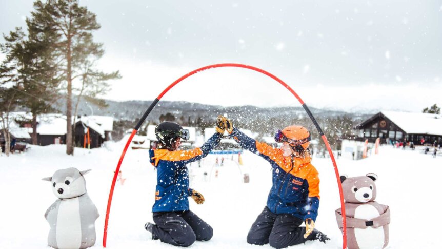 To personer i skiklær gir high-five i snøen, omgitt av snøkledde hytter og vinterpyntede figurer.