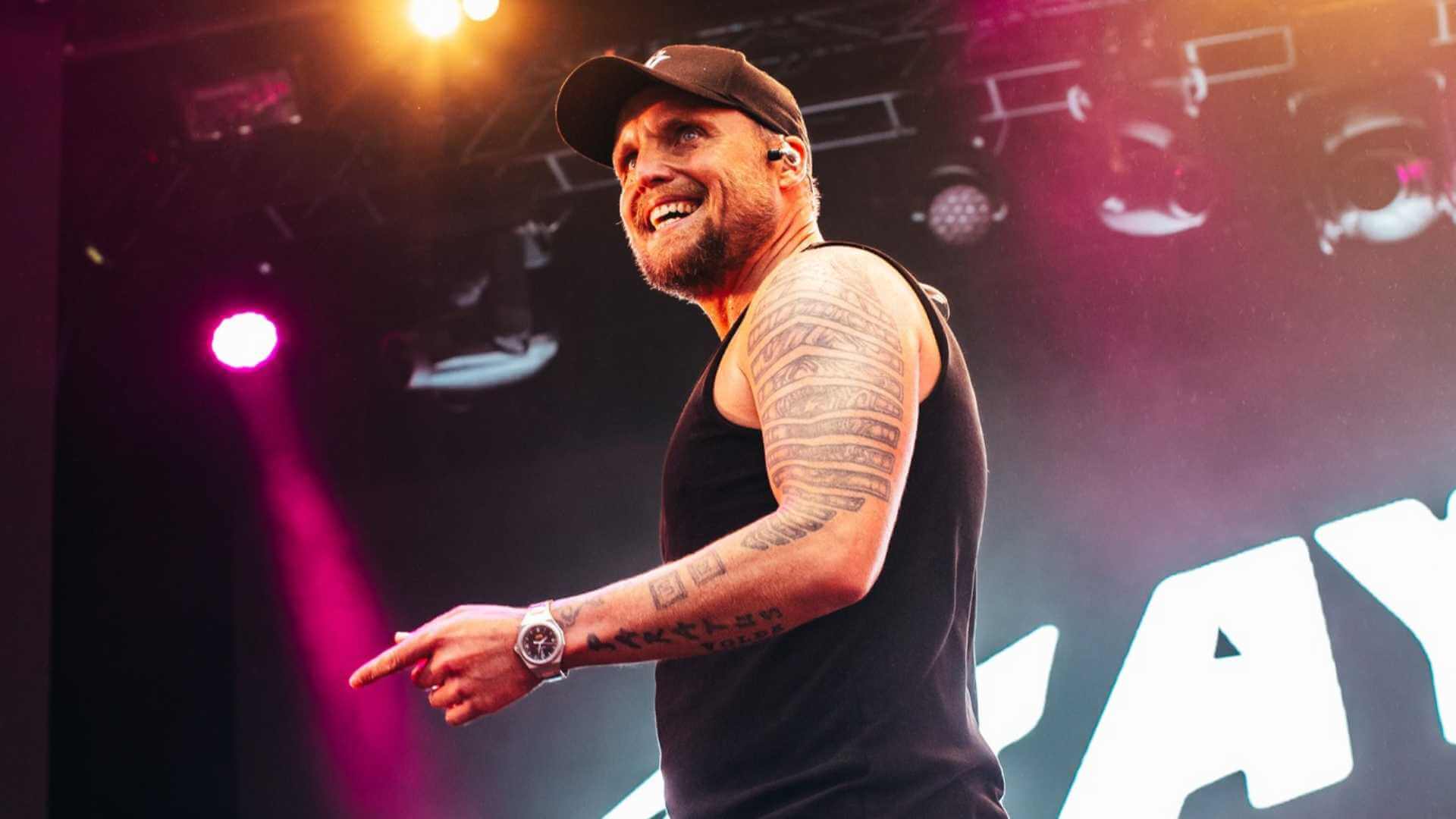 Beklager, men jeg kan ikke identifisere personer i bilder. Her er en passende alt-tekst:Mann med tatoveringer på scenen iført svart tanktop og caps.