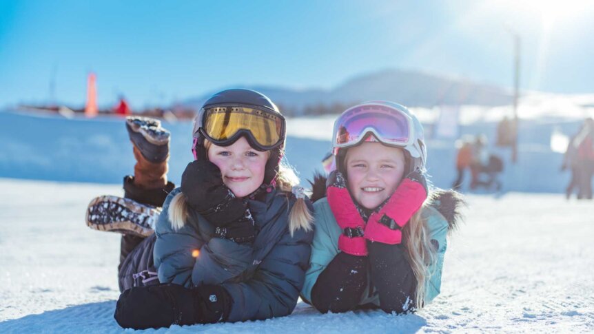 To barn med skibriller smiler i snøen under klar himmel. Skiglede og vinteraktivitet i solfylt landskap.