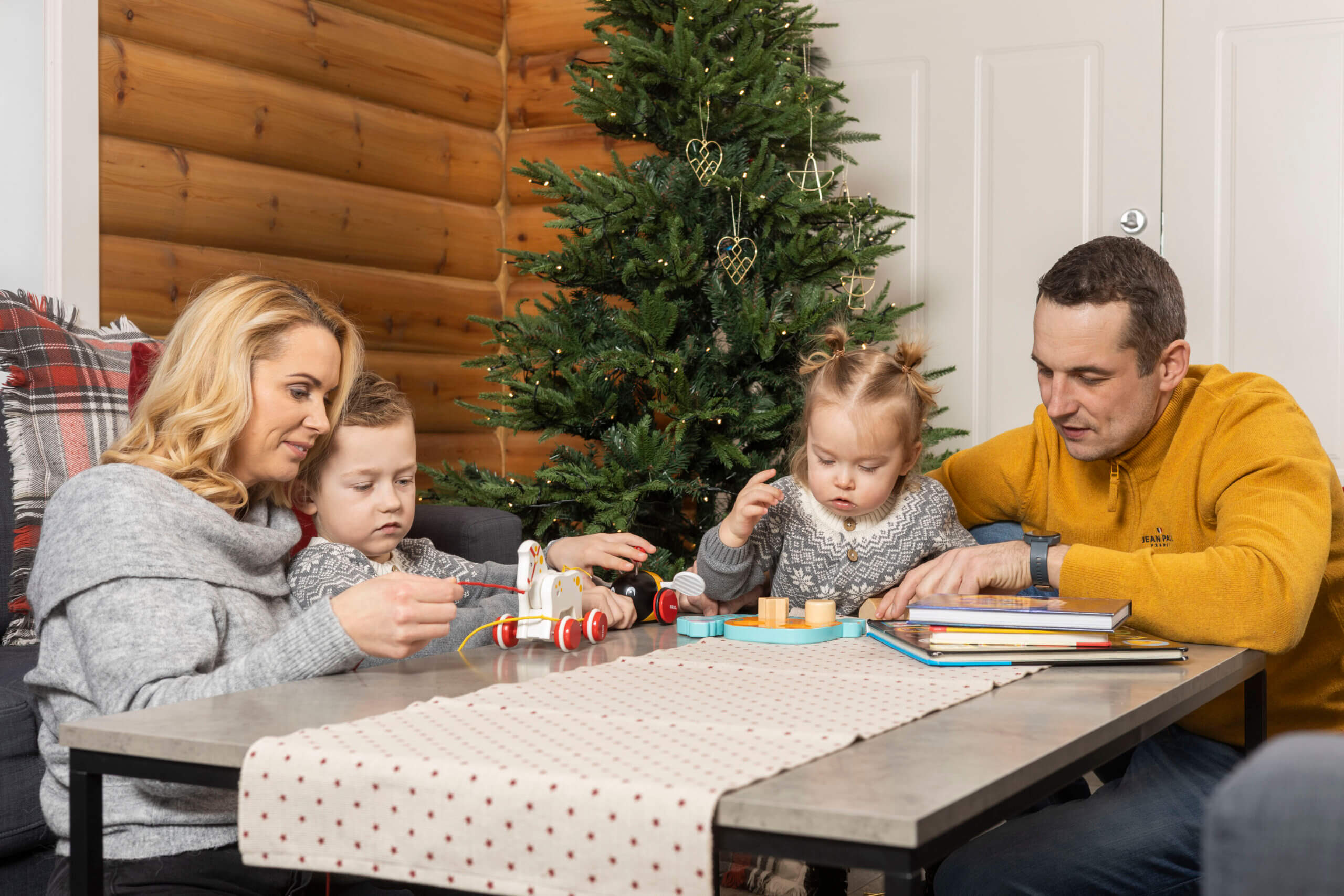 Familie koser seg med leker ved juletreet i stue med trepanelvegger.