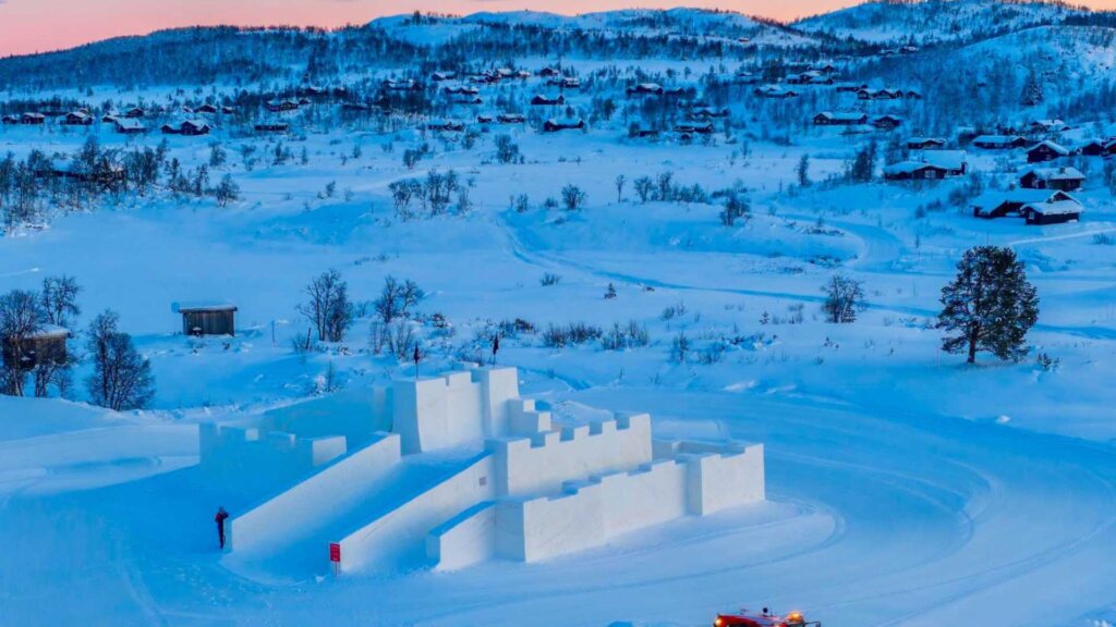 Snøslott i vinterlandskap med fjellhytter i bakgrunnen under solnedgang i Norge.