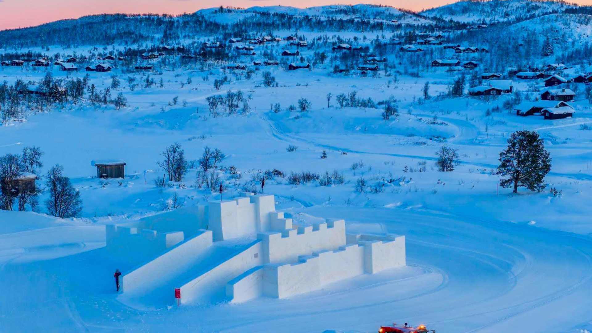Snøslott i vinterlandskap med fjellhytter i bakgrunnen under solnedgang i Norge.