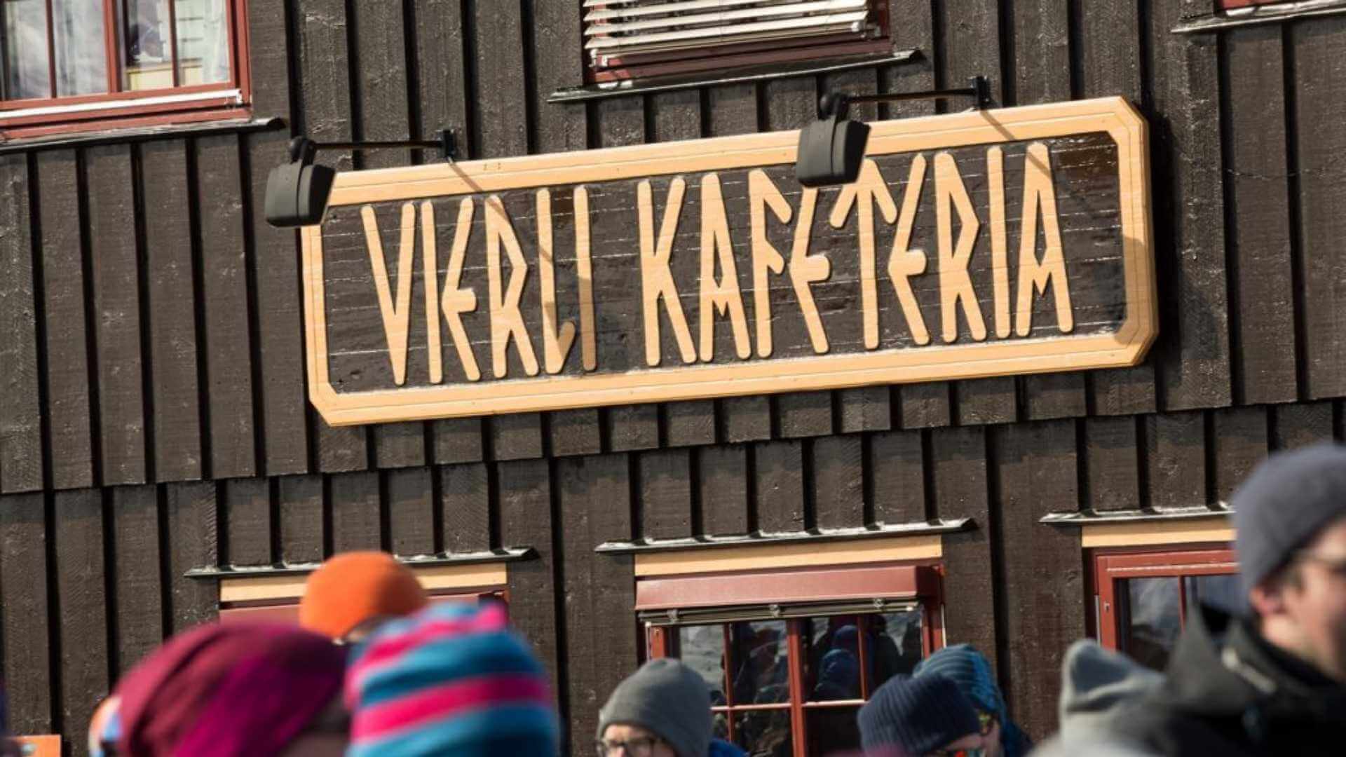 Utvendig skilt for Vekri Kafeteria på en brun trebygning, med folk foran i vinterklær.