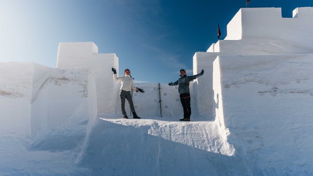 To mennesker poserer i en stor snøborg under klar blå himmel.