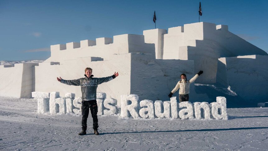 To personer foran en snøborg med This is Rauland i snøtekst, under en klar blå himmel.
