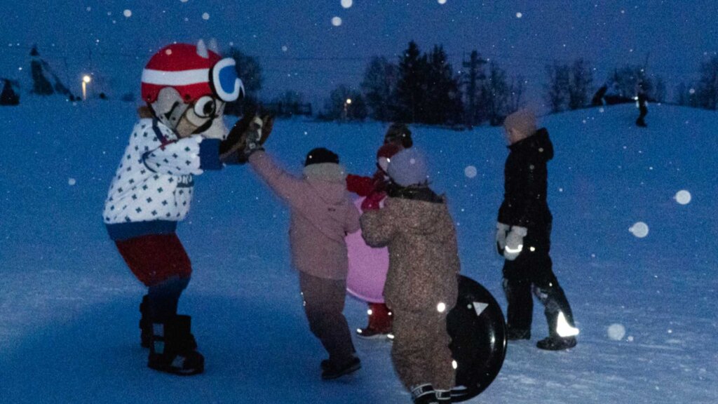 Barn leker i snøen med en maskot i kveldsmørket, iført vinterklær og i leken stemning.