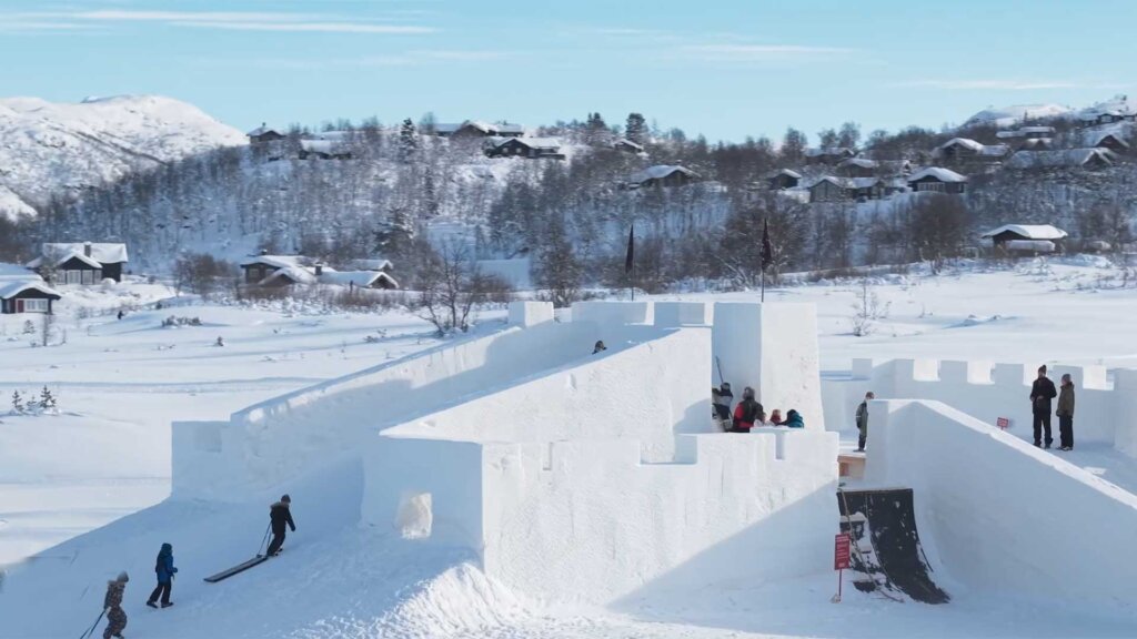 Snøborg i vinterlandskap med barn som leker, fjellhytter og snødekte trær i bakgrunnen.