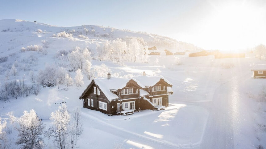 Snødekt norsk fjellandskap med tradisjonell hytte i solskinn, omgitt av vinterkledde trær.