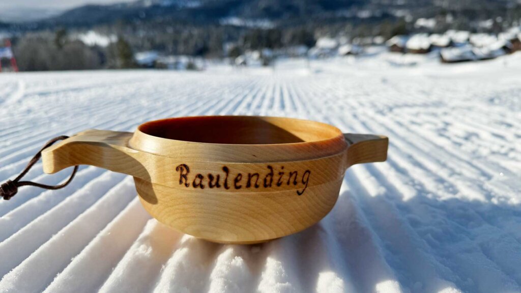 Trehåndlaget kopp med teksten Raulending på snødekt bakgrunn, vinterlandskap i bakgrunnen.
