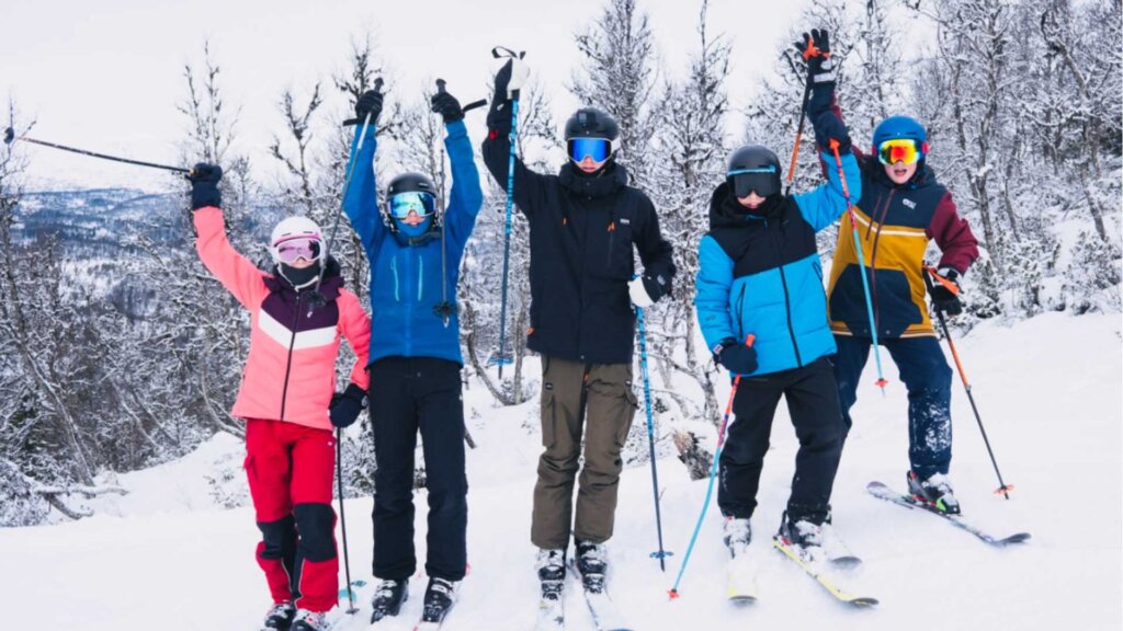 Fem glade skikjørere i snødekte omgivelser, poserer med hevede ski og smilende ansikter.