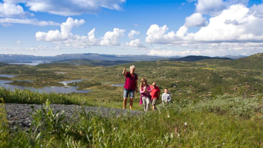 Familie på tur i naturskjønn utsikt med fjell og innsjøer under en blå himmel med skyer.