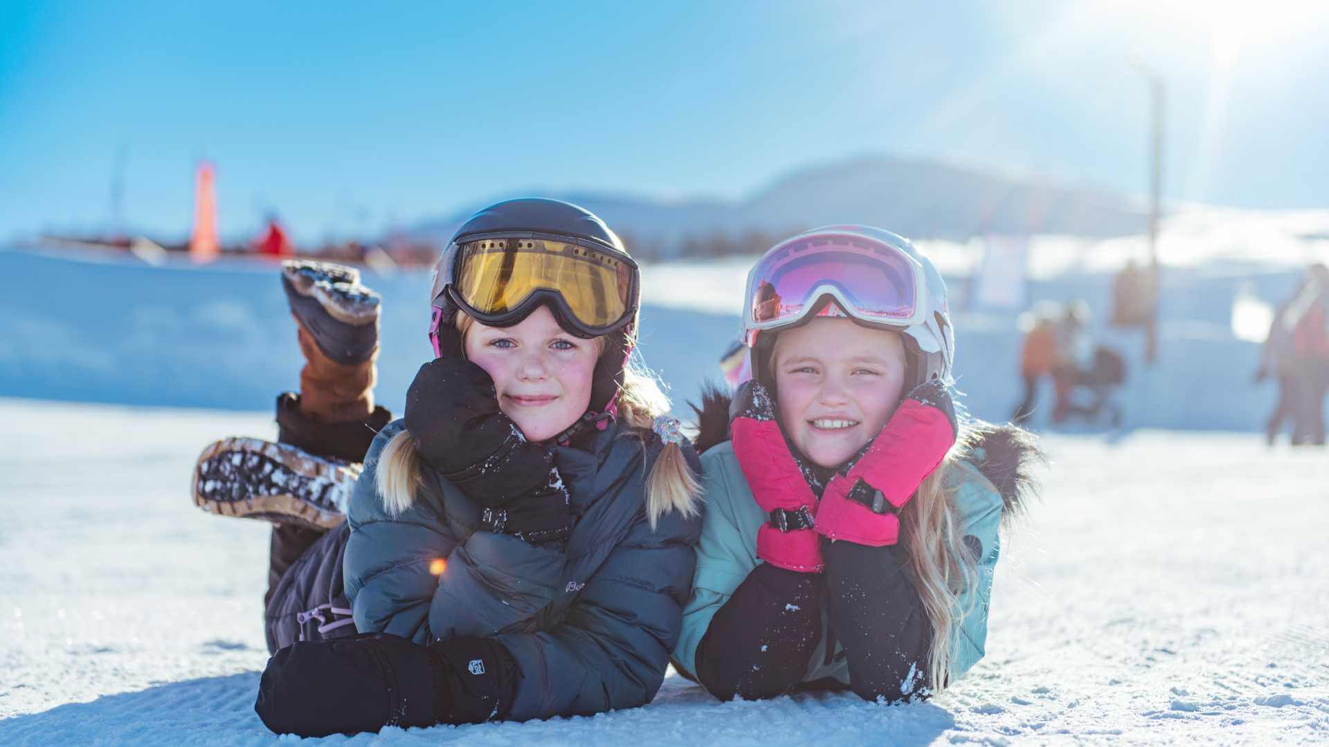 To piger i skitøj ligger smilende på sneen og klar til vintersjov i et solrigt bjerglandskab.