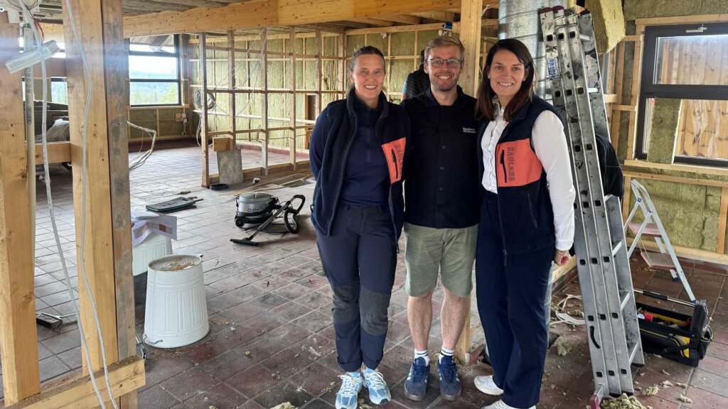 Tre personer poserer i en renovert bygning med byggeutstyr og materialer rundt dem.