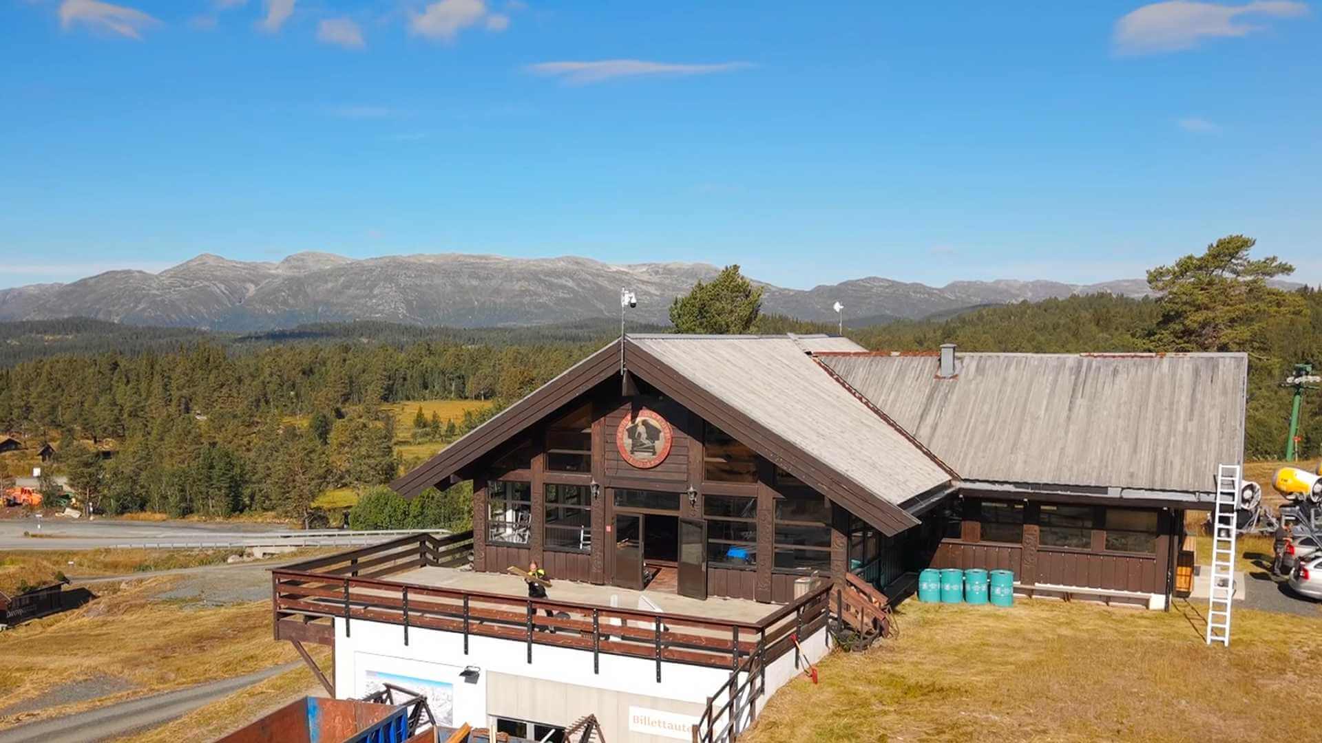 Alt-tekst: Utsikt over tradisjonell hytte med fjellbakgrunn og klar blå himmel i Norge.