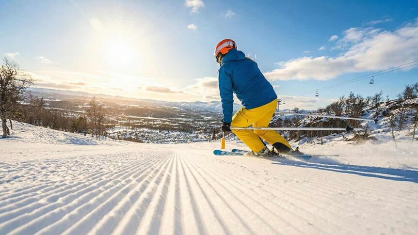 En skiløper i blå jakke og gule bukser nyter solen på en snødekt bakke. Vakkert vinterlandskap i bakgrunnen.