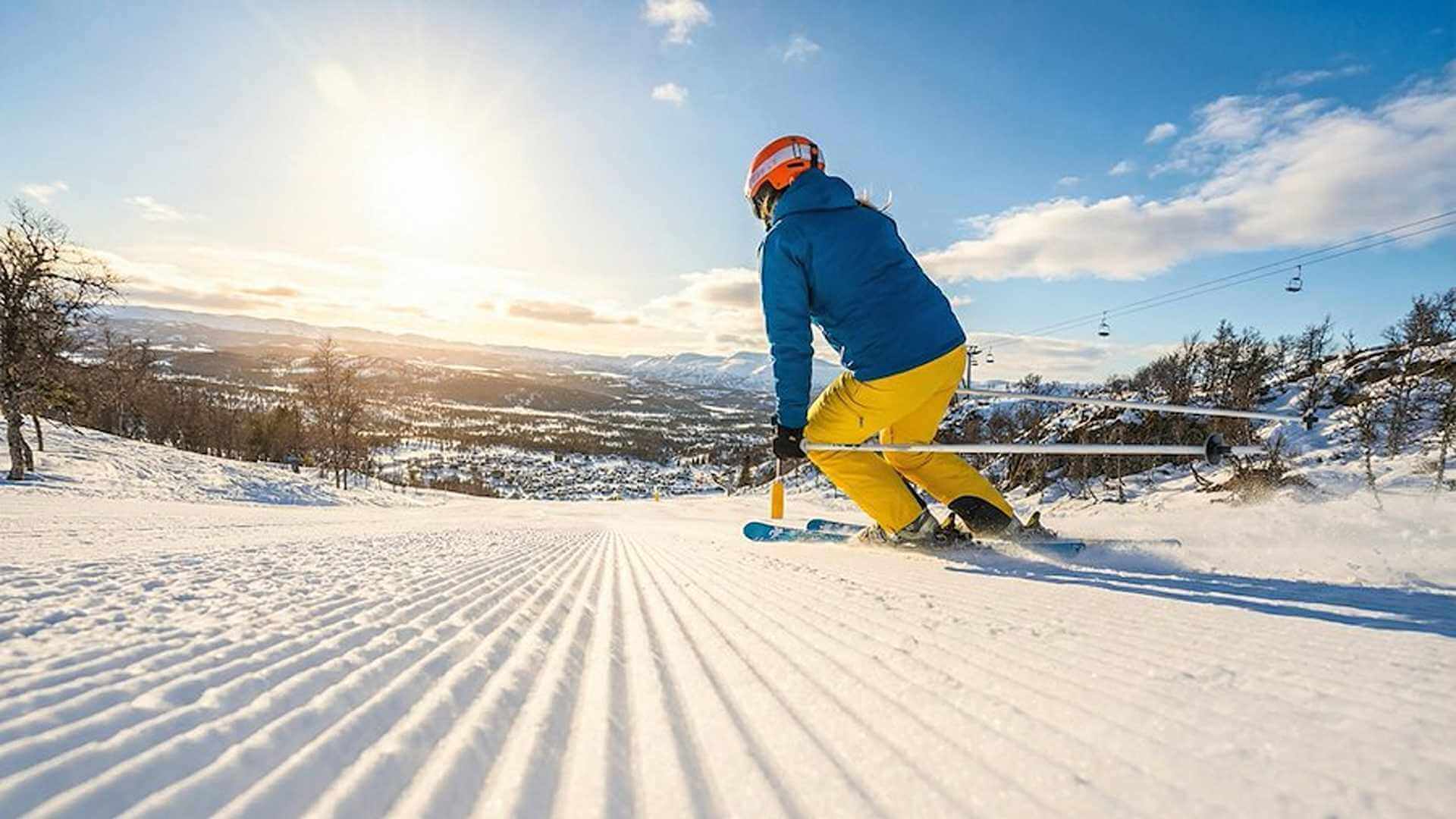 En skiløper i blå jakke og gule bukser nyter solen på en snødekt bakke. Vakkert vinterlandskap i bakgrunnen.