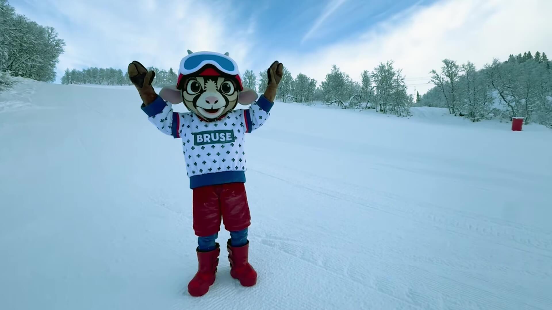 Myskende maskot i sikkerhetsutstyr, som poserer i snødekt skianlegg med klare himmel og trær i bakgrunnen.