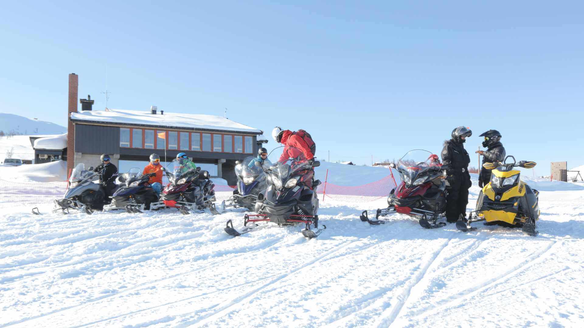 Folk i vinterklær står ved snøscootere foran en hytte i snødekt landskap.