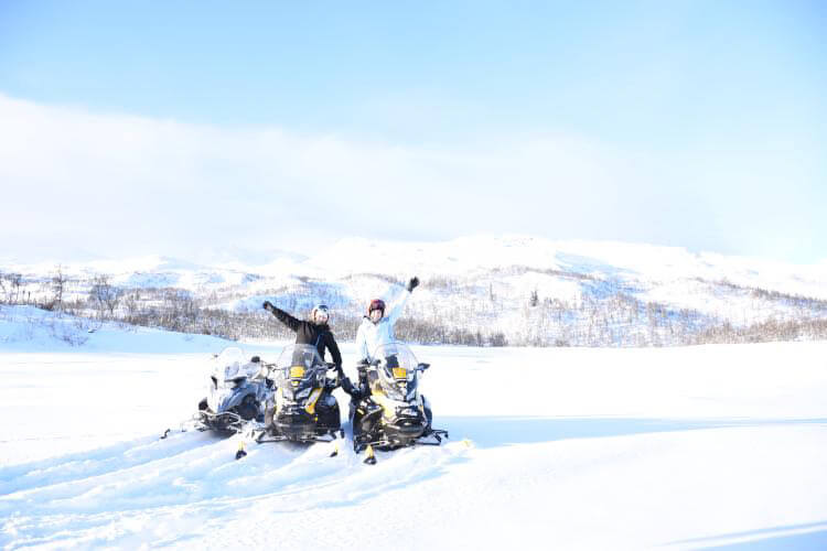 To personer gleder seg på snøskutere i snødekt landskap, med fjell i bakgrunnen under en klar blå himmel.