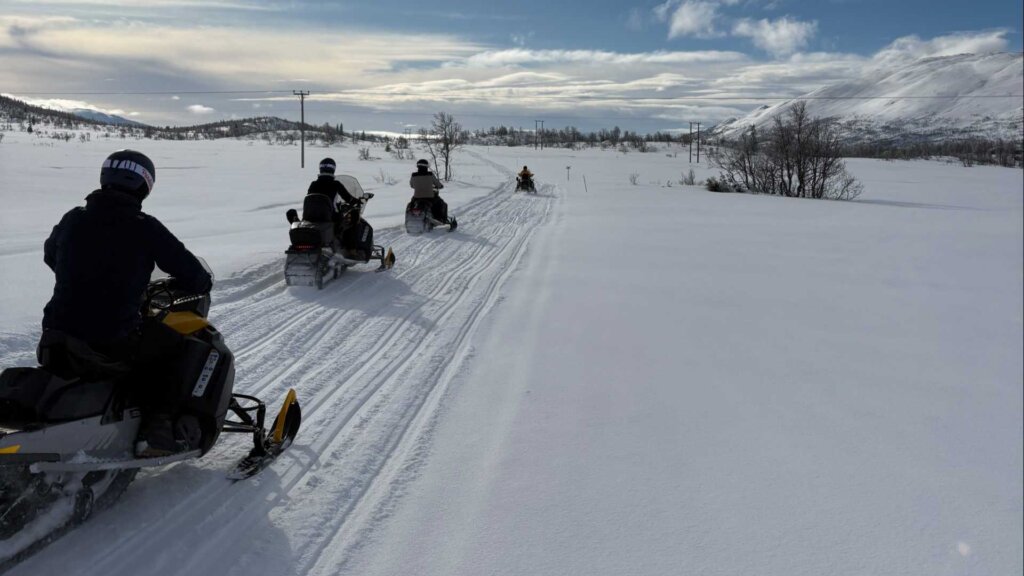 Fire snøskutere kjører gjennom et snødekket landskap under en blå himmel med skyer i bakgrunnen.