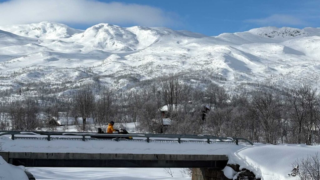 Snøscooterkjører på bro med snødekte fjell og trær i bakgrunnen under en klar blå himmel.