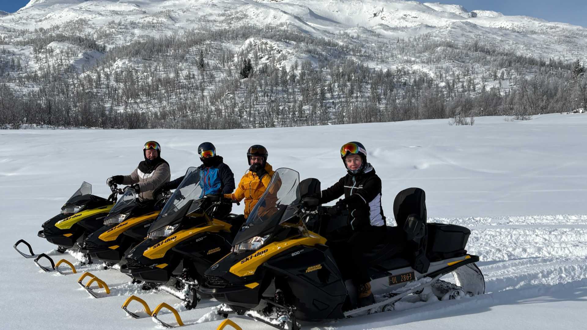 Fire personer på snøskuter i fjellet, omkranset av snødekte landskap og klar blå himmel i Norge.