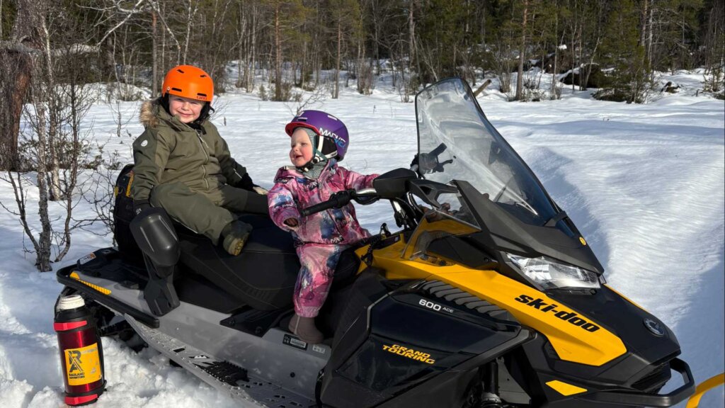 To små barn smiler og kjører scooter i snøen om vinteren, med hjelmer og varme klær.