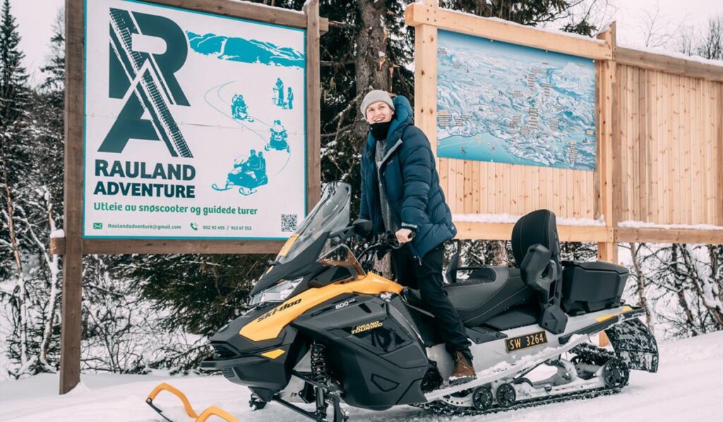En person sitter på en snøscooter ved Rauland Adventure, med snødekte trær og informasjonsplakat i bakgrunnen.