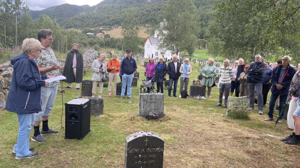 Gruppe mennesker samlet ved en gravplass i naturskjønne omgivelser, lytter til en taler.