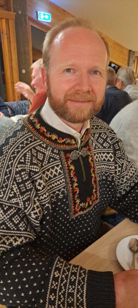 Arnestein Skinnarland, i tradisjonell norsk strikkegenser smiler vennlig ved bord.